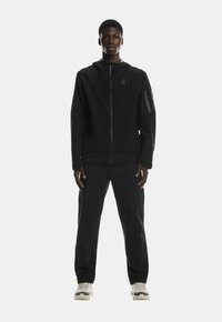 Černá zipová bunda a kalhoty vyrobené z lehkého materiálu, s jemným logem a s kapsou na zip na rukávu. Nošeno s béžovými teniskami.