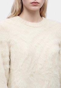 Femme portant un pull en maille duveteuse de couleur crème avec un col rond, montrant le haut de la poitrine et une partie du visage ainsi que des cheveux blonds.
