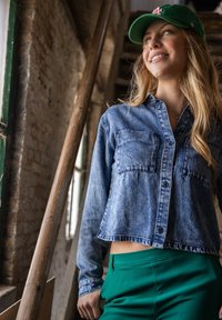 Denim overhemd met knopen, twee borstzakken, korte lengte, gecombineerd met groene broek, voor een rustieke achtergrond.