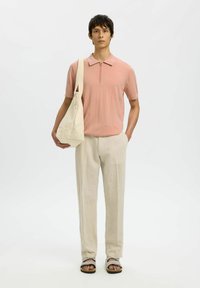 Jeune homme portant un polo à manches courtes rose avec une fermeture éclair, un pantalon en lin beige, des sandales, et portant un grand sac cabas beige sur l'épaule.