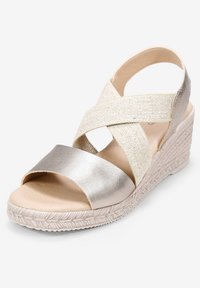Sandalia cuña en tejido metálico plateado y beige. Straps cruzados, diseño de punta abierta y suela texturizada estilo yute. Ligera y transpirable.