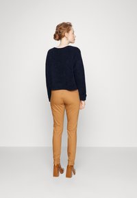 Pull en maille crochet bleu marine, coupe décontractée, col rond. Pantalons ajustés beige, longueur cheville avec une fermeture éclair sur le côté. Chaussures à talons carrés marron.