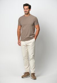 T-shirt marrone a maniche corte, pantaloni color crema e scarpe sportive marroni, in piedi dritto contro uno sfondo neutro.