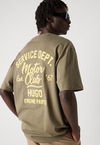Oliven grøn oversized t-shirt med fremtrædende gul grafisk tekst på ryggen, der viser "SERVICE DEPT. Motor Club Est. '67 HUGO ENGINE PARTS."