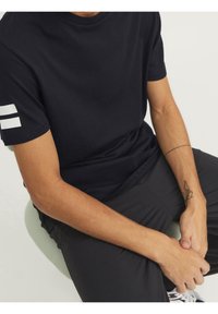 Svart bomullströja med korta ärmar, med två vita horisontella ränder på vänster ärm. Slät textur, avslappnad passform.