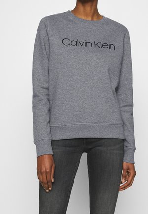 Person iført en grå Calvin Klein sweatshirt og mørkegrå jeans, vist fra nakke til midt på låret med arme afslappet ved siden.