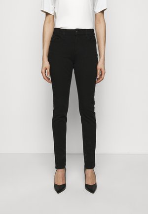 Frau trägt eng anliegende schwarze Hose und schwarze spitze Absätze, steht vor einem schlichten weißen Hintergrund.