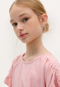Helles rosa T-Shirt mit kurzen Puffärmeln, das zarte Lochstickereien entlang der Schulternähte aufweist. Glatte Baumwolltextur.