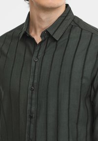 Camisa de botones de color verde oscuro, hecha de una tela suave, con rayas negras verticales, un cuello y botones negros en la parte delantera.