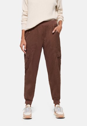 Persona vistiendo pantalones cargo marrones con bolsillos laterales con botones, un suéter crema y zapatillas beige con cordones azul claro sobre un fondo blanco.