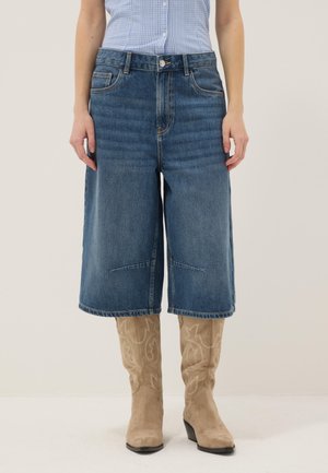 Persona che indossa pantaloncini di jeans blu fino al ginocchio, una camicia a quadri blu infilata dentro e stivali da cowboy beige ricamati, in piedi.