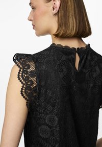 Svart spetsblus med scallopmönstrade ärmar, intrikata mönster, nyckelhålsdetalj på baksidan och texturerad tyg. Elegant och detaljerad design.