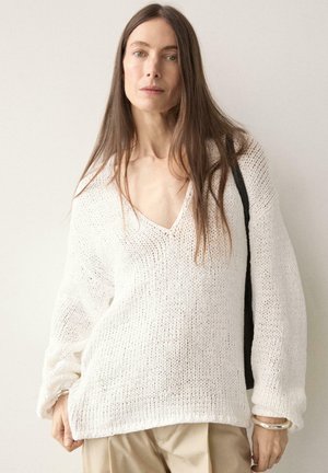 Donna con capelli castani lunghi che indossa un maglione bianco lavorato a maglia ampio e pantaloni beige, con una borsa nera a tracolla su uno sfondo semplice.