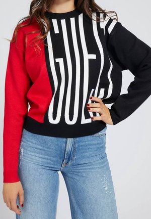 Roter, schwarzer und weißer Strickpullover mit auffälligem "GUES" Schriftzug. Lange Ärmel und gerippte Bündchen mit lässigem Schnitt. Über einer hellblauen Jeans getragen.