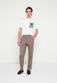 White cotton t-shirt with a green and blue graphic pocket print, paired with light brown tailored trousers and dark brown loafers.