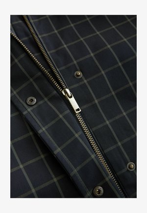 Veste à carreaux navy avec une fermeture éclair dorée, présentant un motif en grille dans des tons de vert foncé et des raccords en laiton. Texture lisse et boutons-pressions visibles.