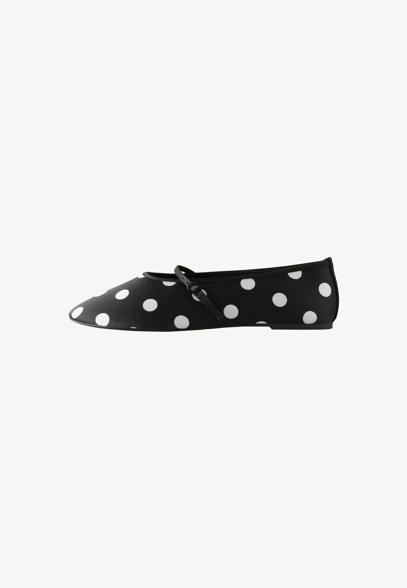 Chaussure plate noire à pois blancs avec une fine lanière traversant le dessus, vue de profil sur un fond blanc.