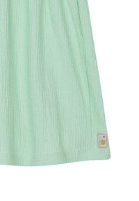 Helles grünes ärmelloses Kleid aus strukturiertem Stoff, mit einem kleinen genähten Etikett mit Obstgrafiken am Saum.