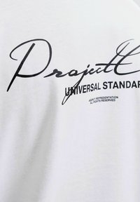 Hvid bomulds t-shirt med stor sort tekst, der læser "Projekt" og "UNIVERSAL STANDARD", med grafiske accenter nedenfor. Glat struktur.