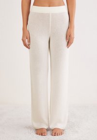 Femme portant un pantalon en tricot large et taille haute blanc, pieds nus sur une surface texturée douce, contre un fond neutre.