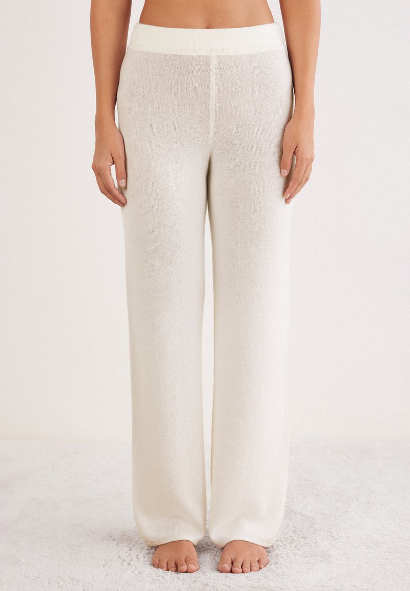 Femme portant un pantalon en tricot large et taille haute blanc, pieds nus sur une surface texturée douce, contre un fond neutre.