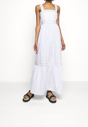 Femme portant une robe maxi blanche sans manches avec des détails cloutés et des sandales plateformes noires, debout devant un fond blanc uni.