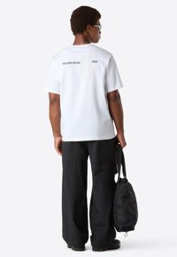 Mand iført en hvid "Never Stop Exploring" t-shirt, sorte brede bukser, sorte sko, holder en sort taske, vender væk mod en ensfarvet baggrund.