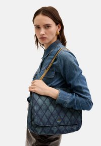 Quiltad denimaxelväska med en guldkedjesnodd, med en slät yta och lockstängning, visar mot en neutral bakgrund.
