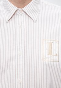 Chemise blanche à boutons avec fines rayures verticales beige et un écusson brodé beige avec la lettre « L » sur la poitrine.