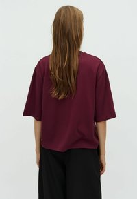 Burgundy T-shirt with a relaxed fit, short sleeves, and a smooth texture, paired with loose black pants, viewed from the back.