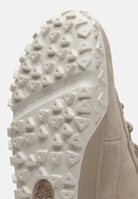 Semelle de sneaker beige clair en caoutchouc texturé, présentant un motif triangulaire pour l'adhérence. Le côté est lisse avec un détail subtil de logo.