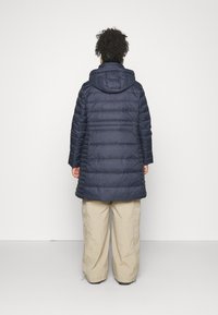 Marineblauer Steppmantel mit Kapuze, mit gesteppter Optik und mittellangem Schnitt. Getragen über hellfarbenen, weit geschnittenen Hosen.