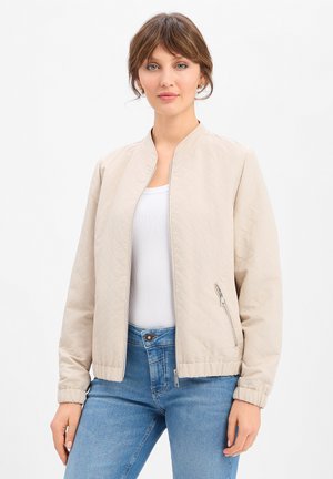 Vrouw draagt een beige doorgestikte bomberjas, wit geribd tanktopje en blauwe spijkerbroek, staand tegen een effen witte achtergrond.