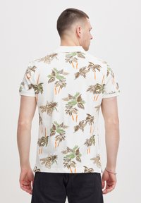 Hvit polo skjorte med et livlig palme mønster i grønn, brun og oransje. Korte ermer, myk tekstur og klassisk passform.