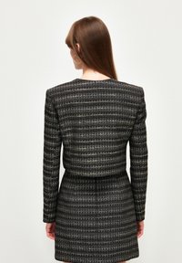 Vestido chaqueta tejido en negro con hilo metálico dorado. Diseño estructurado, mangas largas y cintura ajustada. Acabado texturizado.