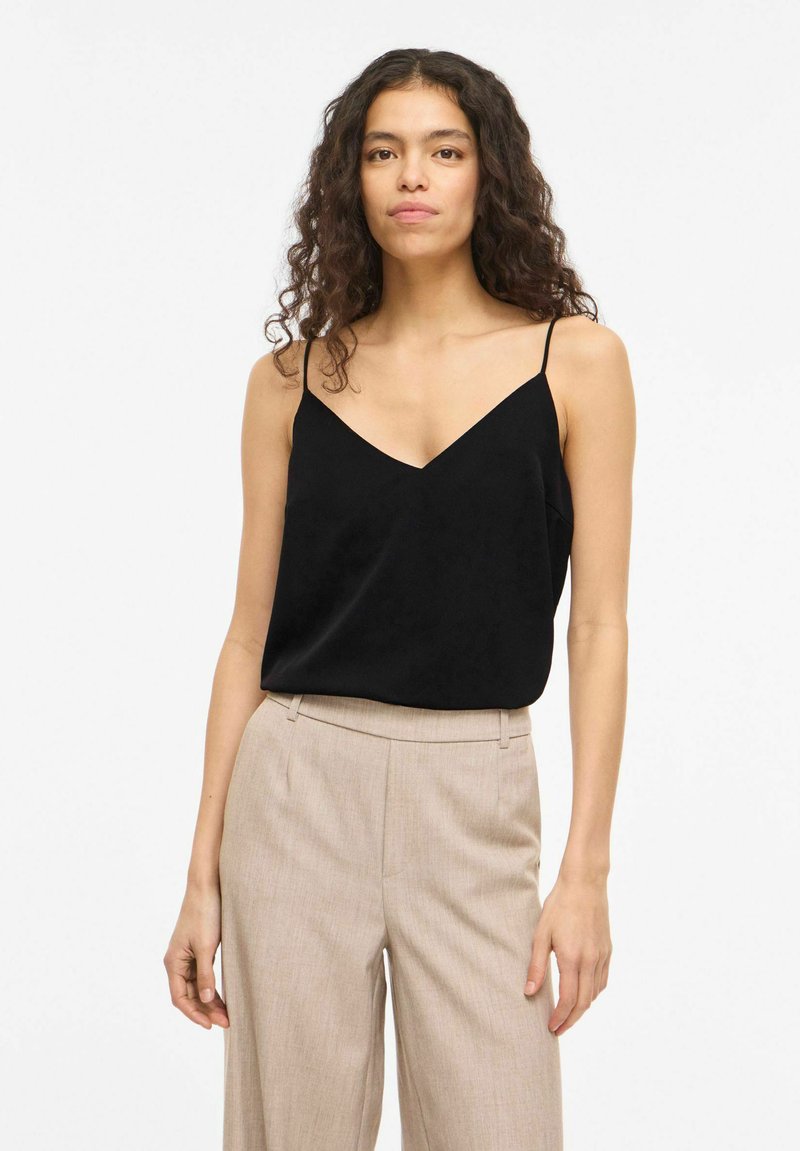 Kvinde med langt krøllet hår iført en sort, ærmeløs V-hals top og beige højttaljede bukser, stående foran en ensfarvet baggrund.