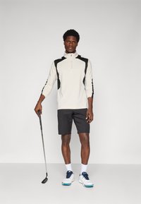 Lys beige langermet polo med svarte detaljer, halv-glidelås i halsen, kombinert med svarte shorts og hvite golfsko. Holder en golfkølle.