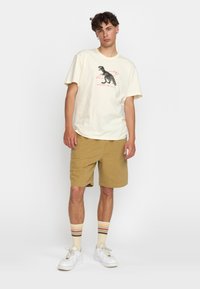 Crème t-shirt met een grafisch ontwerp van een dinosaurus, beige shorts en witte sneakers met meerkleurige gestreepte sokken. Casual pasvorm en lichtgewicht materiaal.