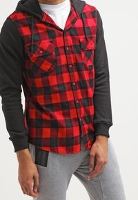 Camicia in flanella a quadri rossi e neri con cappuccio, dotata di chiusura con bottoni e due tasche sul petto, abbinata a pantaloni della tuta grigi.