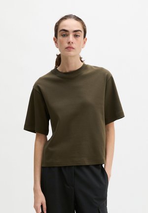 Femme aux cheveux châtains attachés portant un t-shirt à manches courtes vert olive et un pantalon noir sur un fond clair uni.