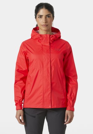 Kobieta w jasnoczerwonej wodoodpornej kurtce z kapturem, wyposażonej w kieszenie na zamek, i czarnych spodniach, stojąca na jednolitym jasnym tle.