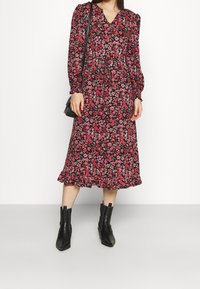 Robe midi fleurie avec un fond noir, ornée de fleurs roses et vertes, manches bouffantes, encolure en V et ourlet à volants. Portée avec des bottines noires.