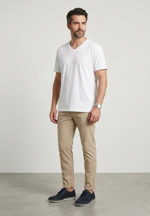 Mand stående iført en hvid kortærmet V-hals shirt, beige bukser, mørkeblå sko og et armbåndsur mod en ensartet lys baggrund.
