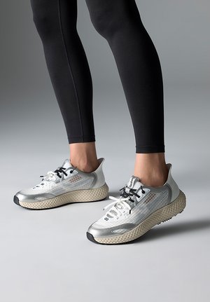 Person wearing silver and beige athletic sneakers with black and white laces, standing on a gray surface in black leggings.