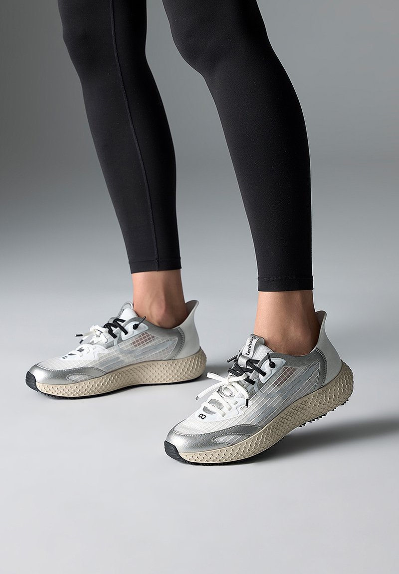 Person iført sølv- og beige atletiksneakers med sorte og hvide snørebånd, stående på en grå overflade iført sorte leggings.