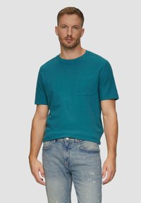 Turkusowa koszulka z krótkim rękawem i okrągłym dekoltem, z kieszonką na lewej piersi, w połączeniu z jasnoniebieskimi dżinsami w stylu distressed.