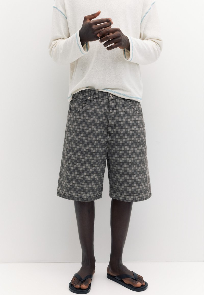 Personne portant une chemise crème à manches longues, un short gris à carreaux arrivant aux genoux et des tongs noires, debout contre un mur blanc.