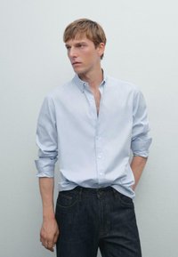 Jonge man met opgerolde mouwen draagt een lichtblauw overhemd met knopen en donkerblauwe jeans, staande tegen een effen lichtgrijze achtergrond.