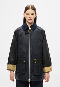 Tengerészkék viaszos dzseki kordbársony gallérral, arany színű fémrészletekkel és két nagy első zsebbel; kockás részletek a mandzsettákon.