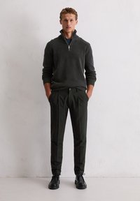 Donkergrijze ritsvest, groene broek en zwarte schoenen. Het vest heeft geribbelde mouwen en zoom, de broek heeft een rechte pasvorm en een knoopsluiting.